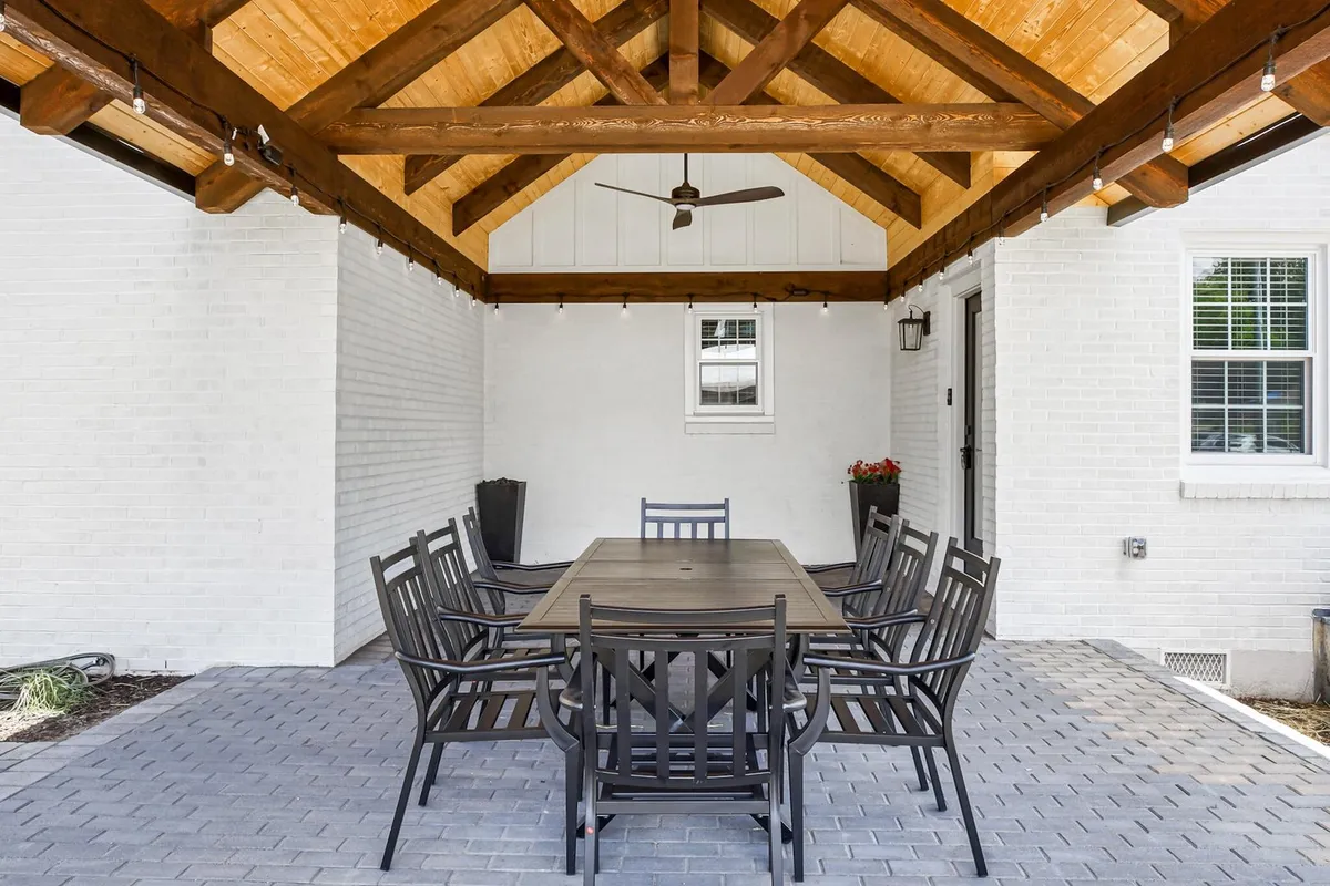 A spacious outdoor dining and lounge area under wooden beams with a ceiling fan, surrounded by greenery. The setting includes a large dining table and comfortable seating, evoking a welcoming atmosphere for both relaxation and social gatherings.