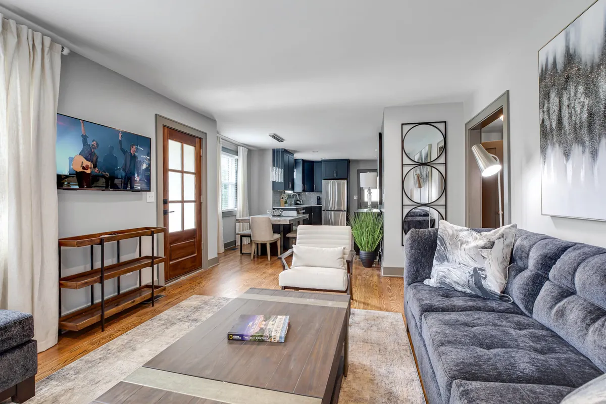 The image features a bright, modern living space with a cozy sofa, inviting coffee table, stylish artwork, and an open view into the dining area and kitchen entrance, all accented by natural light and thoughtful decor.