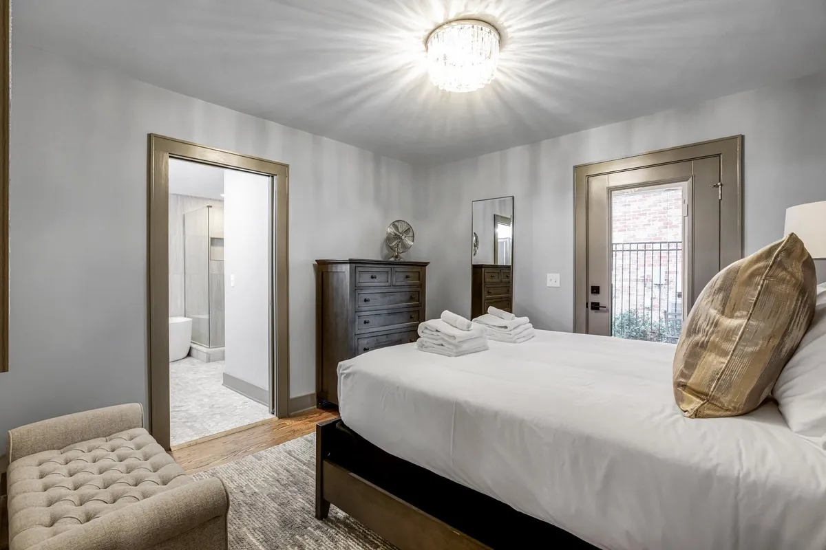 A comfortable bedroom with a queen bed topped with plush pillows, a dresser for ample storage, a charming chandelier, and a door that opens to an outdoor patio. Sunlight streams in, creating a welcoming and soothing space.