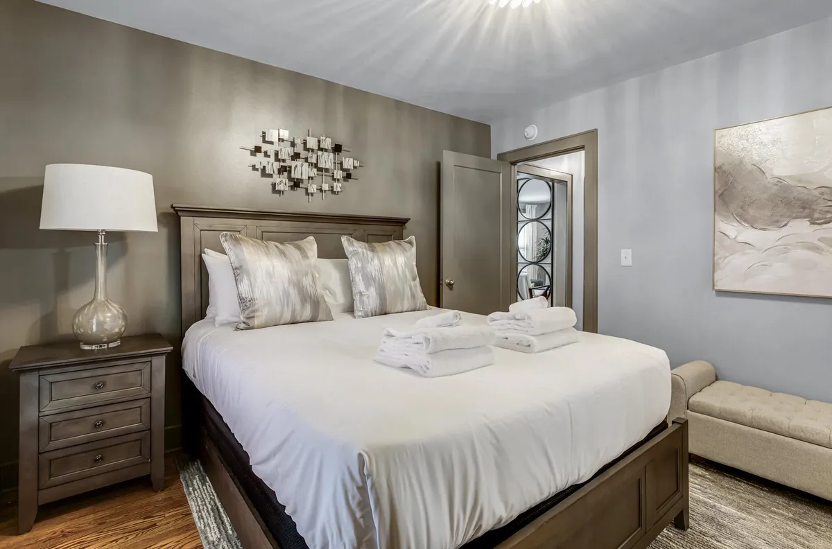 A tranquil bedroom featuring a plush queen bed adorned with decorative pillows, modern wall art above the headboard, and a nightstand with a softly glowing lamp. Gentle natural light filters in, enhancing the serene and cozy atmosphere.