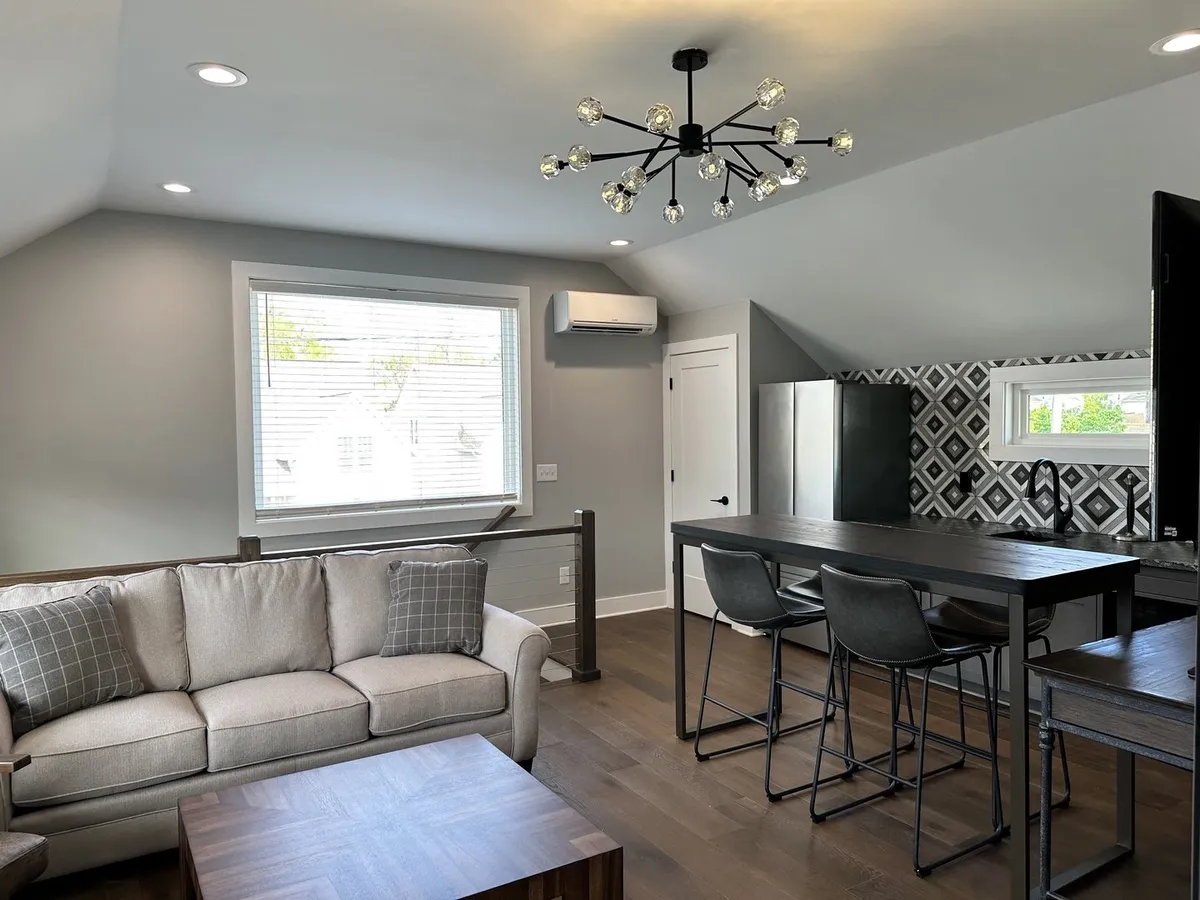A chic living area with a sofa adorned with decorative pillows, a geometric wallpaper accent wall, a modern chandelier, and a dining table with stools. The space blends areas for relaxation, entertaining, and dining in a visually striking setting.