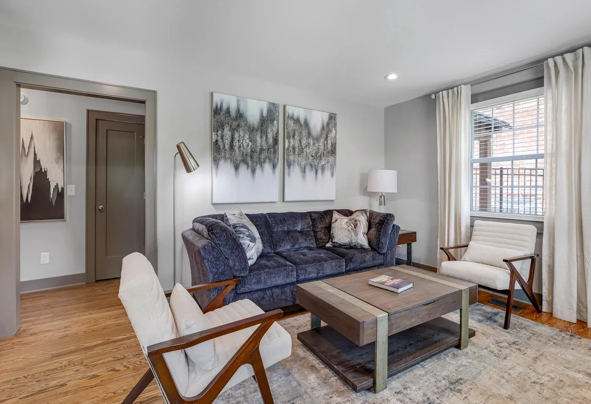A cozy living area featuring a comfortable sofa, eye-catching artwork on the wall, a coffee table, and a floor lamp. The room is well-lit and inviting, creating a space ideal for relaxing, reading, or socializing.