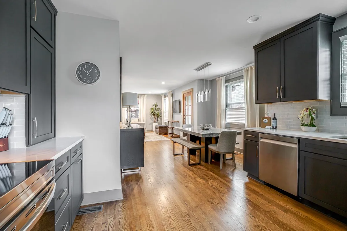 A view of a kitchen and dining area filled with natural light, showcasing a kitchen island, large dining table, plants, and decorative wall elements. The space feels welcoming and is arranged to support both cooking and shared dining experiences.