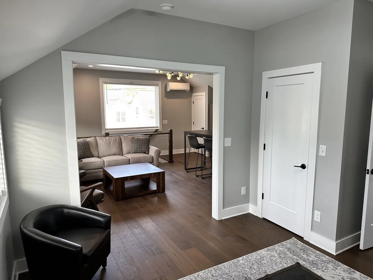 A bright, inviting sitting area with a comfortable couch, wooden coffee table, black armchair, and large window letting in natural light. The setup supports both relaxation and productivity—ideal for morning coffee, reading, or working remotely.