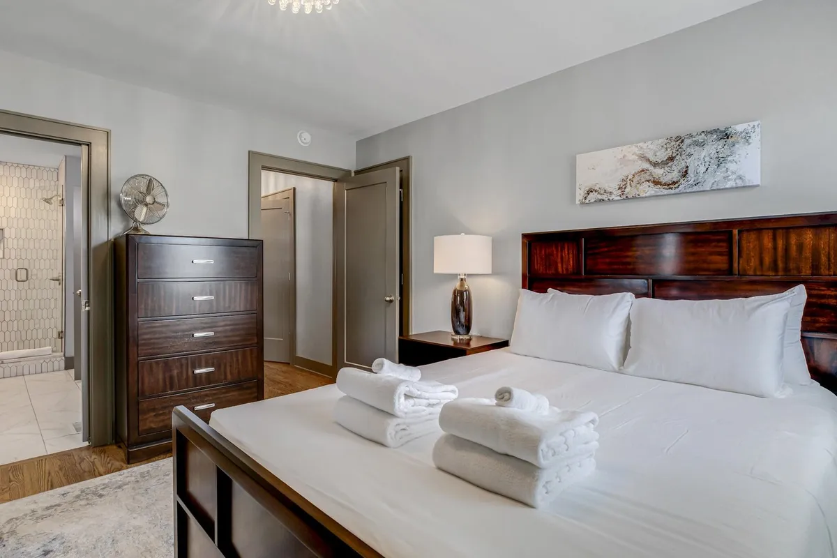 A serene bedroom scene with a neatly made bed, towels arranged on top, a nightstand with a lamp, dresser, and artwork on the wall. The mood is calm and restful, perfect for unwinding after a day in Nashville.